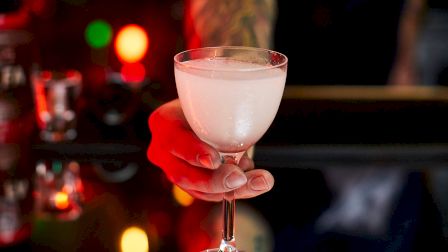 A person is handing a frothy white cocktail in a glass, with colorful bokeh lights in the background creating a festive atmosphere.