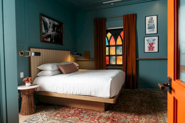 A cozy bedroom with teal walls, a bed, colorful window, and artwork; features a rug, wooden accents, and orange curtains and door.