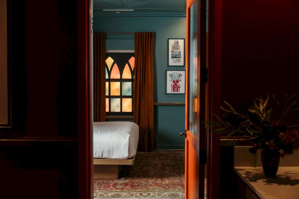 A room with a bed, colorful stained glass window, curtains, art on walls, and a rug, seen through an open door.