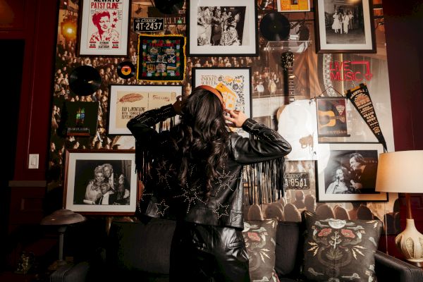 A person in a leather jacket faces a wall filled with framed photos, posters, and music memorabilia, adding a vintage vibe to the room.
