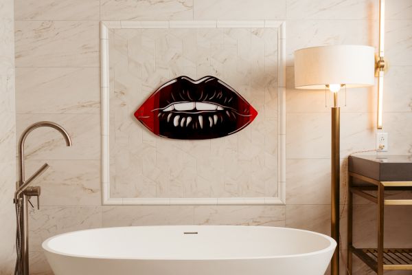 A modern bathroom with a white freestanding tub, artwork of lips on the wall, and stylish lighting and furniture accents complete the look.