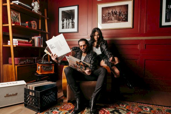 A man and woman are sitting in a vintage-style room with records and a record player, surrounded by books and framed photos on the wall.