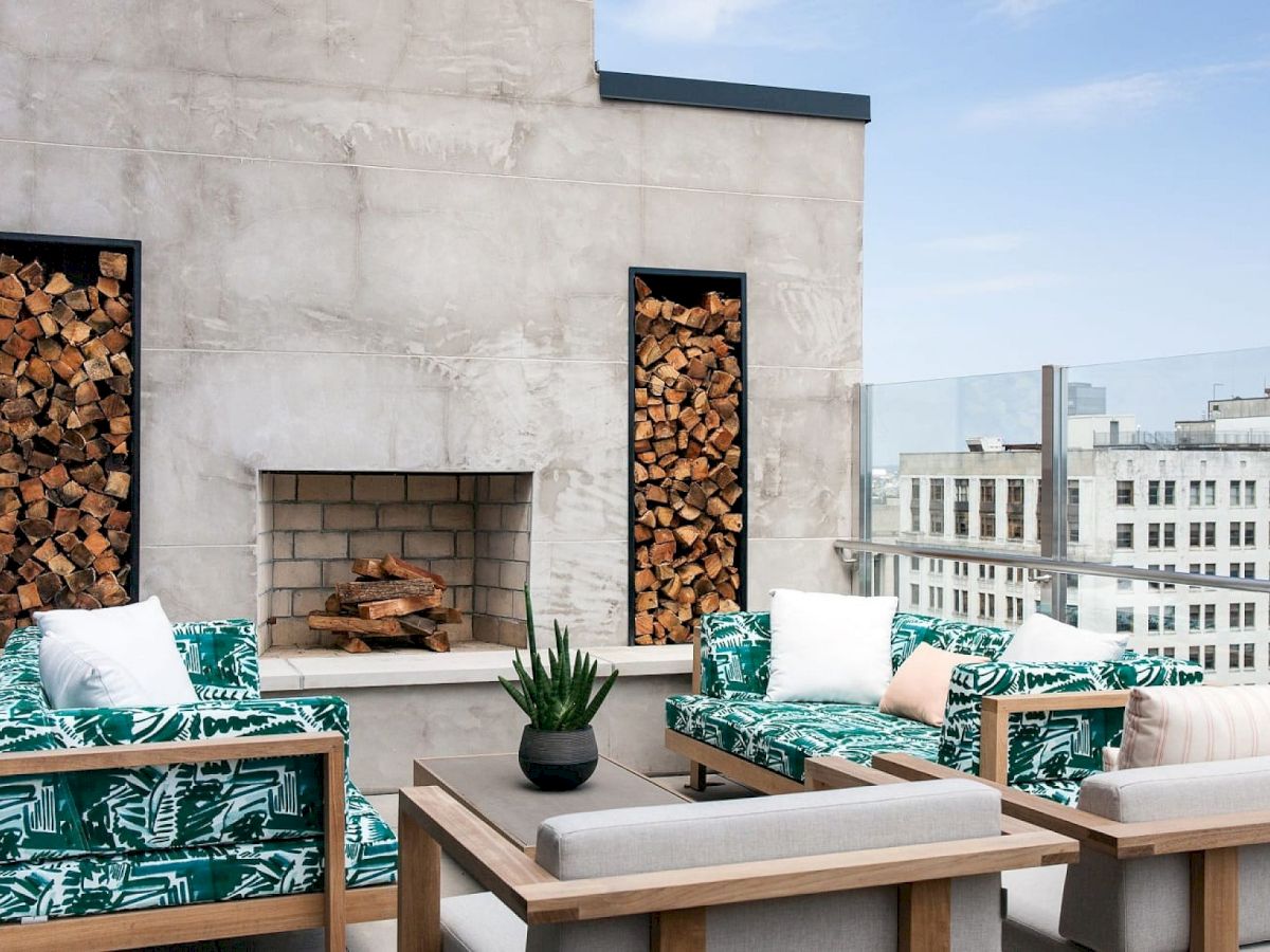 A rooftop patio with sofas, a small plant, and decorative wood panels by a fireplace, overlooking city buildings under a blue sky.