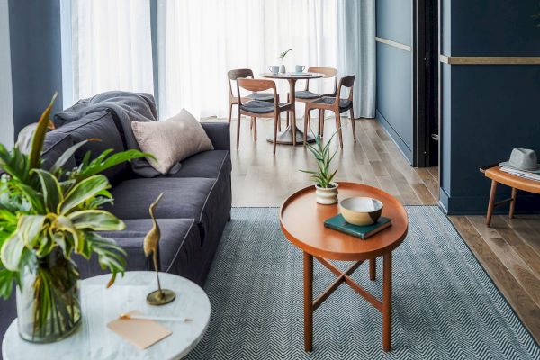A modern living room with a gray sofa, plants, a round wooden coffee table, and a dining area with chairs in the background.