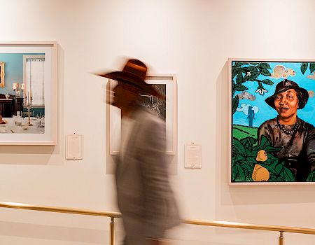 A person in a hat walks past colorful framed art on a gallery wall.