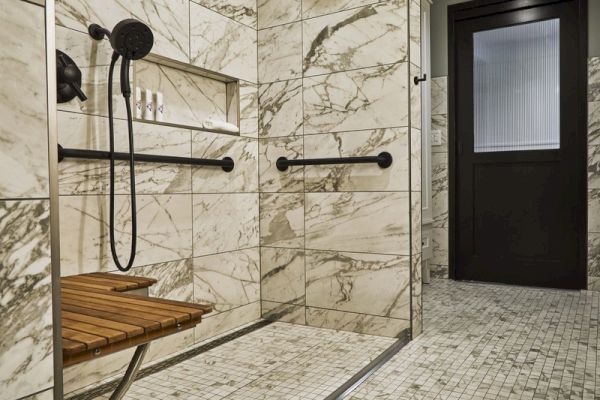 The image shows a luxurious bathroom with marble walls, a wooden bench, a handheld showerhead, and grab bars.