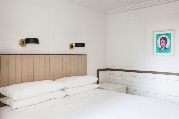 A minimalist bedroom features a bed with white linens, two wall lamps, and a framed pop art portrait on the wall.