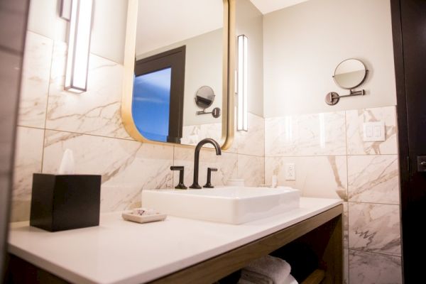 A modern bathroom with a marble countertop, white sink, black faucet, illuminated mirror, tissue box, and round wall-mounted mirror.