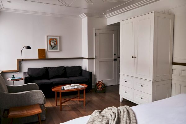 A cozy room with a sofa, lamp, armchair, coffee table, framed artwork, door, and wardrobe. Light walls and wood flooring complete the look.