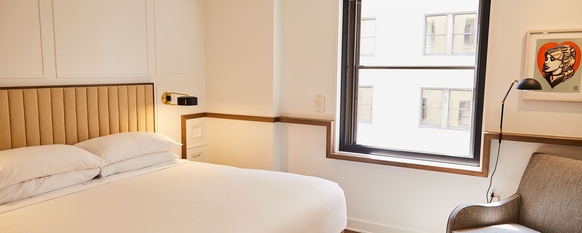 A modern hotel room with a bed, armchair, lamp, and window.