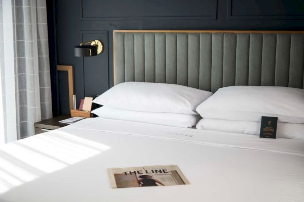 A neat bed with a gray upholstered headboard, white pillows, bedside table, wall lamp, and a magazine titled 