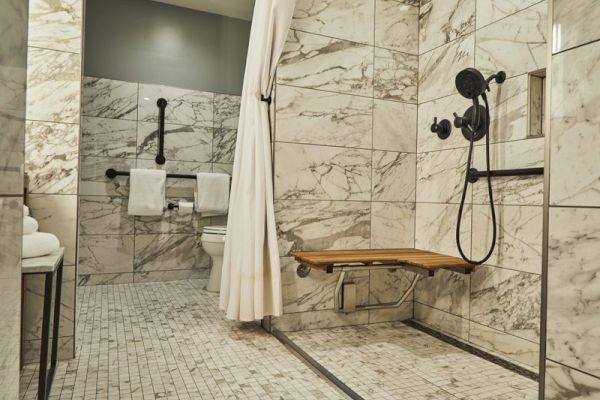 A restroom with marble tiles, a shower with a bench, handheld showerhead, curtain, towels on racks, and grab bars near the toilet.