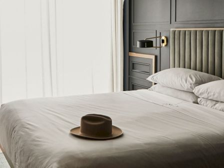 A neatly made bed with a hat on top, near a window with curtains.