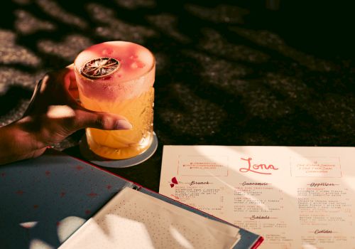 A hand holds a drink with a slice of dried fruit beside an open menu.