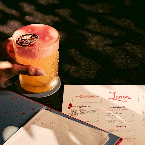 A hand holds a drink with a slice of dried fruit beside an open menu.