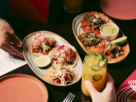 Tacos on plates with lime, a drink with jalapeño slices, and a hand holding a menu.