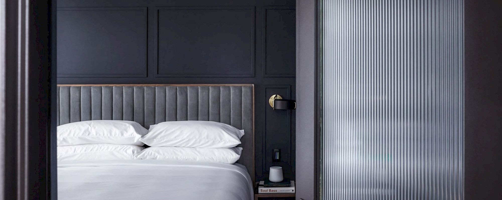 A modern bedroom with a gray headboard and white bedding seen through a glass-paneled door.