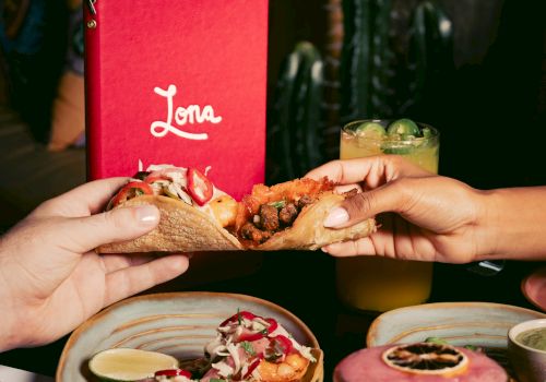 Two hands holding tacos, with a red menu and drinks in the background.