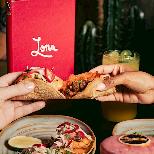 Two hands holding tacos, with a red menu and drinks in the background.