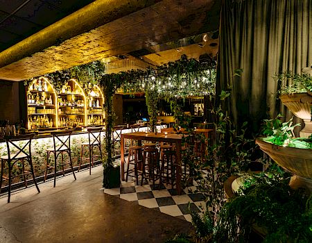 A warmly lit bar with wooden stools, greenery, and a decorative fountain.
